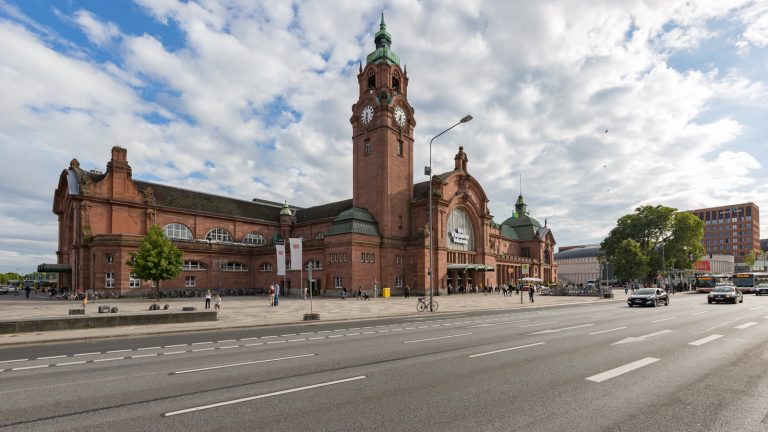 Der historische Kopfbau des Wiesbadener Hauptbahnhofs aus rotem Sandstein mit hohem Uhrturm, davor eine breite mehrspurige Straße.