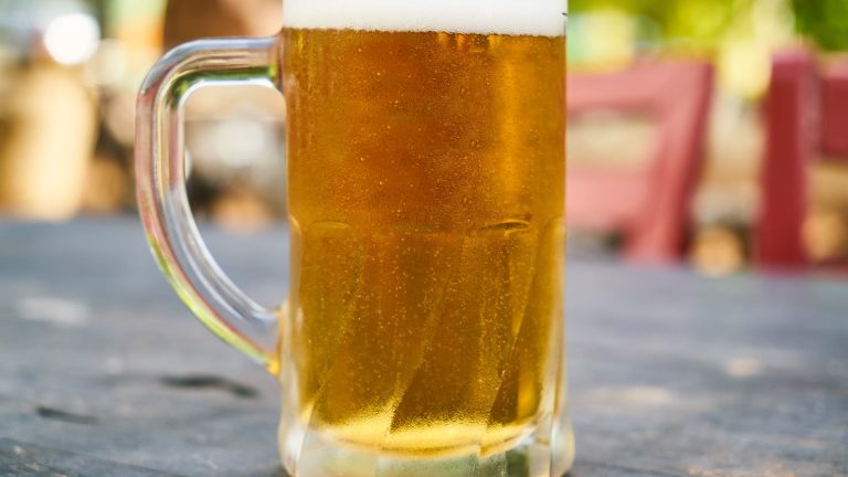 Ein beschlagener Bierkrug mit heller Füllung und Schaumkrone steht auf einem verwitterten Holztisch im Freien, im unscharfen Hintergrund rote Sitzbänke.
