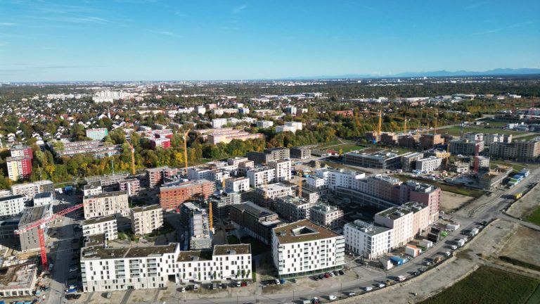 Luftaufnahme eines neuen Stadtviertels am Stadtrand: helle mehrgeschossige Wohnblöcke mit Flachdächern, dazwischen Baukräne und noch unbebaute Flächen, am Horizont die Alpen.