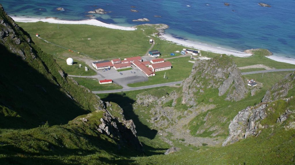 Luftaufnahme des Raketengeländes von Andøya Space in Nordnorwegen: niedrige rot-weiße Gebäude und eine weiße Radarkuppel auf einer grünen Landzunge zwischen felsigen Hängen und einem hellen Sandstrand am blauen Meer.