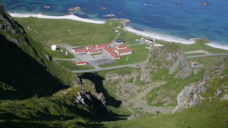 Luftaufnahme des Raketengeländes von Andøya Space in Nordnorwegen: niedrige rot-weiße Gebäude und eine weiße Radarkuppel auf einer grünen Landzunge zwischen felsigen Hängen und einem hellen Sandstrand am blauen Meer.